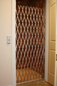 A residential elevator with a metal accordion-style gate partially closed, revealing a wooden interior and a control button mounted on the wall outside.