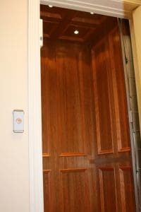 A home elevator with polished wooden panel walls, recessed ceiling lights, and a white exterior doorframe. A small round elevator button is mounted on the wall outside the elevator.