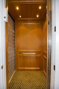 A small residential elevator with wooden paneled walls, a patterned carpet floor, ceiling lights, and a metal accordion gate partially open on the left side.