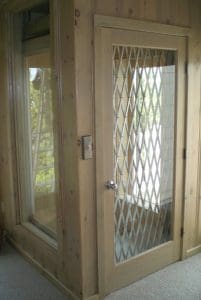 A small residential elevator with a wooden frame and a door featuring diamond-patterned metal bars, next to a matching window, both set in light-colored wood paneling.