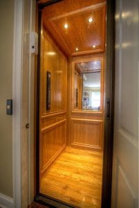 A small residential elevator with wood-paneled walls, a wood floor, ceiling lights, a control panel, and a large mirror at the back, viewed from an open doorway.
