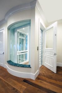 A home elevator with glass walls and a white door, located in a modern interior with wood flooring and cream-colored walls. The elevator is next to a curved window alcove with a blue-green ledge.