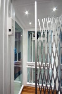 A modern home elevator with glass walls, bright ceiling lights, a folding metal gate, and wooden flooring. The elevator appears compact and sleek, with a control panel on the wall.