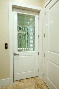 White home elevator door with a glass panel showing a metal accordion safety gate inside. The elevator is located next to another closed door, and there is a button panel on the wall. Light-colored tile flooring is visible.