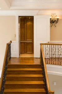 A wooden staircase with a black iron railing leads to a hallway, where an open white door reveals a wooden-paneled elevator. A decorative wall sconce is mounted on the beige wall to the right.