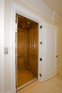 A residential elevator with wooden interior and metal control panel is shown behind an open white door in a home hallway.