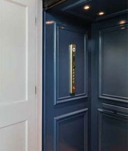 A modern home elevator with glossy dark blue paneled walls, a control panel with buttons on one side, and ceiling lights, located next to a white door.
