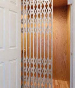 A small wooden-paneled residential elevator with an accordion-style metal safety gate partially open, next to a white door.