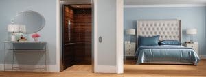 A modern bedroom with light blue walls, a large bed with a tufted headboard, two nightstands, and a console table with a round mirror. An open residential elevator with wood paneling is built into the room.