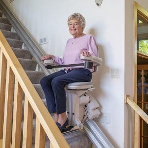 An older woman in a pink top and navy pants smiles while riding a stair lift chair up a carpeted staircase, showcasing the comfort and convenience of residential straight stair lifts inside her home.