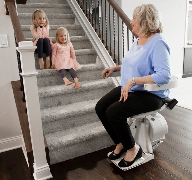 Woman seated on a Access Stair Lift ascending stairs with two smiling granddaughters nearby in a cozy home setting.