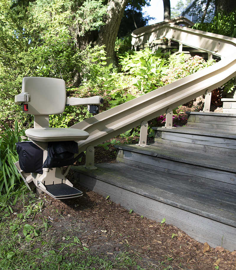 Bruno Elite Outdoor Curved Stairlift on Wood Deck With Plants in Background, With Lift at Bottom of Rail