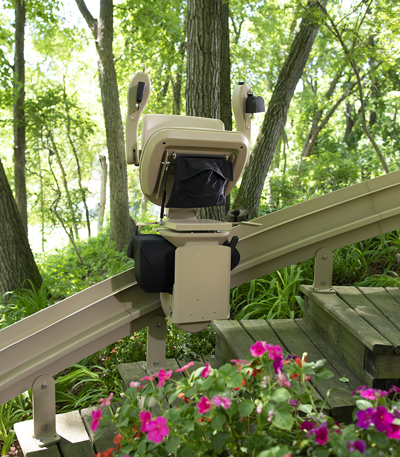 Bruno Elite Heavy Duty Outdoor Curved Stair Lift with Flowers in Front and Trees in Back. Center of Rail in Folded Position