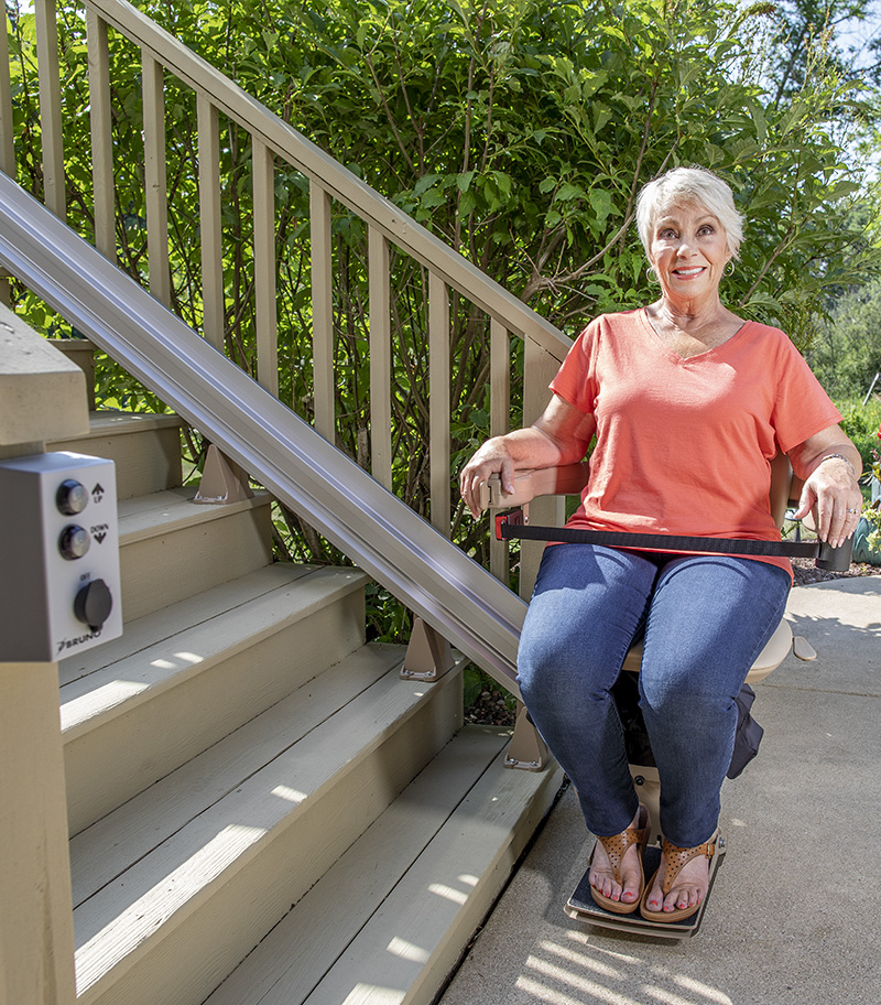Bruno Elite Straight Bariatric Straight Outdoor Stairlift with Rider at Bottom of Stairs with Plants and Fixed RF Call-Send