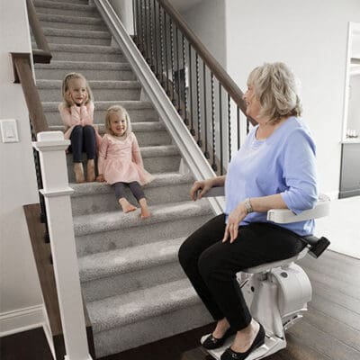 An woman sits on a stair lift at the base of a carpeted staircase, smiling at two young girls sitting together on the bottom steps, all appearing happy and relaxed in a modern home.