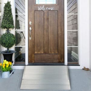 A front door with “Welcome” and glass panels is approached via a National Ramp Aluminum Threshold Ramp over a light concrete porch. On the left are a potted topiary and yellow tulips.