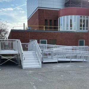A building entrance has stairs and a Commercial Ramp, both metal with railings, leading to a brick building with large windows. The Commercial Ramp is ADA compliant and designed for accessibility.