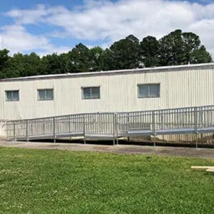 A portable classroom features a Commercial Ramp at its entrance, placed on a grassy area surrounded by trees, with a partly cloudy sky overhead.
