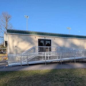 A portable classroom with green trim features a Commercial Ramp for wheelchair access at the front. Trees and sports field lights stand in the background beneath a clear blue sky.