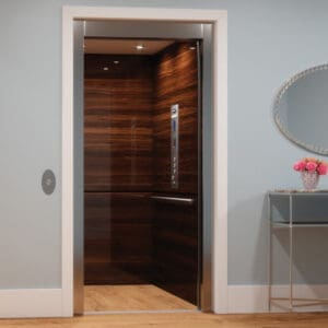 An Eclipse Elevator by Savaria with wood-paneled walls and stainless steel trim stands open. Next to it, a silver console table holds a small vase of pink flowers beneath a round mirror. The flooring is light wood.