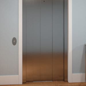 Closed silver Eclipse Elevator doors by Savaria set in a white frame within a light blue wall. A round elevator button is on the left, while part of a radiator appears to the right and brown wooden flooring lies below.
