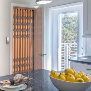 A modern kitchen showcases a bowl of lemons on a dark countertop. In the background, the Luxury Laminated Technology Elevator by REI, with an accordion-style gate and open door, adds elegance as sunlight streams through a large window.