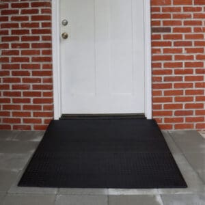 A black Rubber Threshold Ramp by National Ramp is positioned in front of a white two-panel door with brass hardware, set in a red brick wall and surrounded by gray tile flooring.
