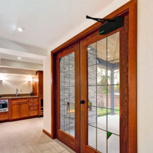 Double glass French doors with wooden frames, fitted with an Automatic Door Opener by Open Sesame above, separate the kitchen and living area from the patio. The kitchen features wooden cabinets and modern appliances.