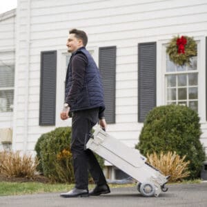 A man in dark clothing pulls a folded Tri-Fold Portable Wheelchair Ramp by National Ramp along a driveway outside a white house with black shutters and a holiday wreath on the window.