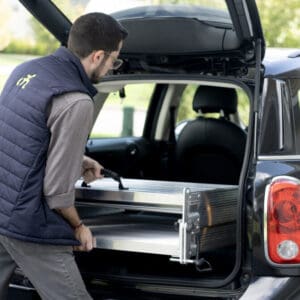A man in glasses and a navy vest lifts a Tri-Fold Portable Wheelchair Ramp by National Ramp into the open trunk of a black car outdoors during the day.