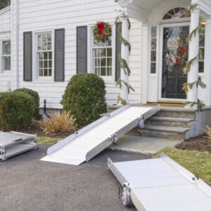 A Tri-Fold Portable Wheelchair Ramp by National Ramp leads up the front steps to a white house adorned with garland and a holiday wreath. Extra ramp sections nearby display this temporary accessibility solution.