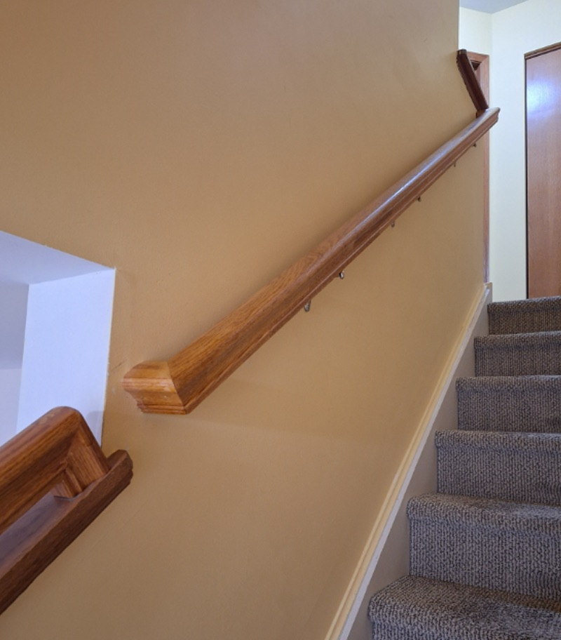 Wooden stair handrail installed along a carpeted residential staircase with beige walls, providing added support and stability for navigating stairs while complementing the home's interior.