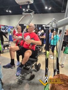 A man in a red shirt is lifted by a mechanical patient lift while seated in a wheelchair at an indoor expo. A smiling woman stands beside him, and people are seen in the background near display booths. The ACCESS logo appears in the corner.
