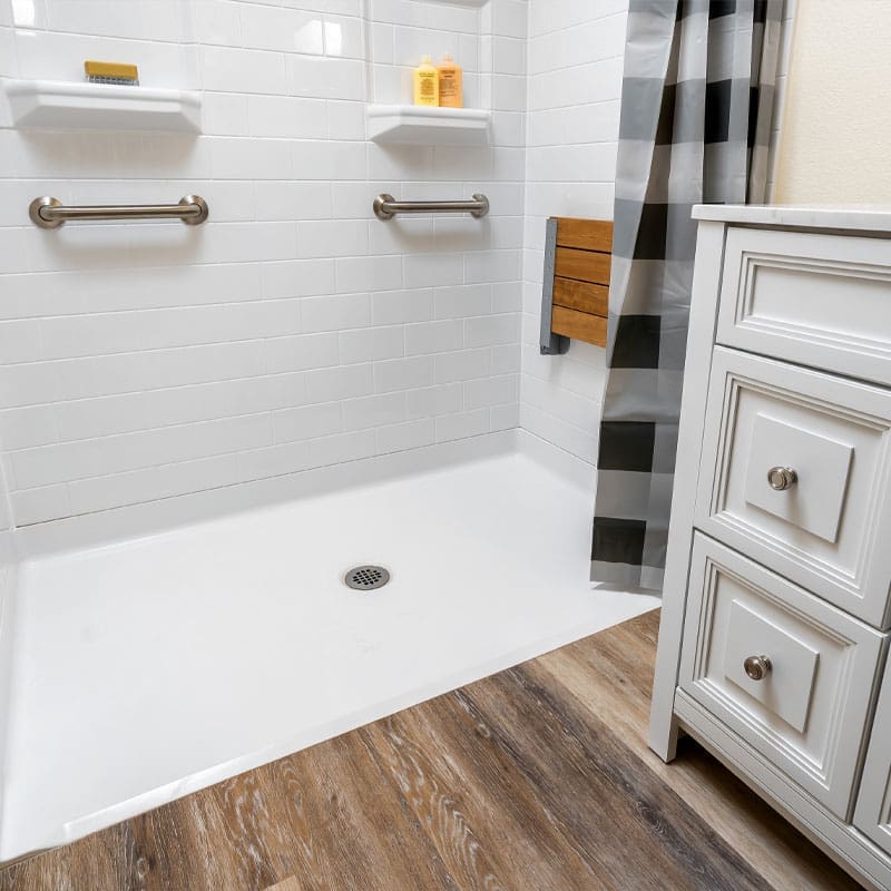 The Roll-In Shower by Best Bath features white tile walls, two grab bars, built-in shelves with toiletries, a fold-down wooden seat, a gray and white striped shower curtain, wood-look flooring, and sits beside a white bathroom vanity. The Roll-In Shower by Best Bath features white tile walls, two grab bars, built-in shelves with toiletries, a fold-down wooden seat, a gray and white striped shower curtain, wood-look flooring, and sits beside a white bathroom vanity.
