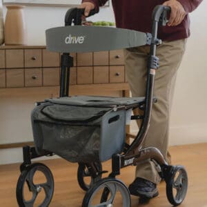 A person pushes a black and gray Drive Nitro rollator walker, an ADA-compliant mobility aid with large wheels and a storage pouch, indoors on a wooden floor.