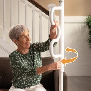 An older woman with short gray hair in a green polka dot blouse installs a white Wonder Pole by Stander next to a couch, showing how to twist and adjust it, with an arrow indicating the motion.