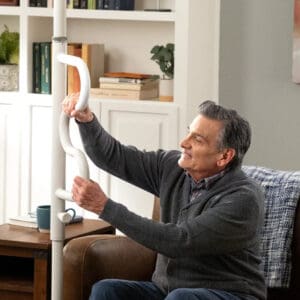 An older man in a gray sweater sits on a couch, smiling as he holds the Wonder Pole by Stander with handles in his living room. Shelves filled with books and decor add warmth to the background.