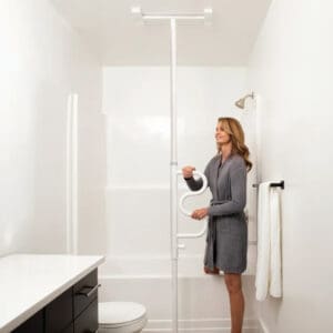 A woman in a gray robe stands in a white bathroom, holding the Wonder Pole by Stander with a curved handle next to the bathtub and toilet for added stability and support.
