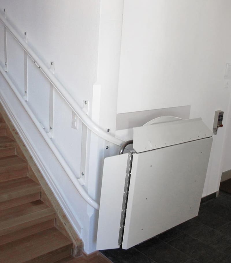 The Pegasus Inclined Platform Lift by Savaria is installed beside a staircase with white railings. Designed for wheelchair users, the platform neatly folds up against the wall when not in use.