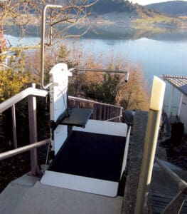 A Pegasus Inclined Platform Lift by Savaria is installed on outdoor stairs beside a lake with trees and hills in the background, offering commercial accessibility up and down the steep staircase.