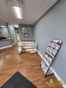 A modern office with wood floors, gray walls, a stairlift demo on a short set of stairs, framed photos, a magazine rack, and the Access Elevator Decatur logo in the corner—highlighting solutions like home elevators and wheelchair ramps in Illinois.