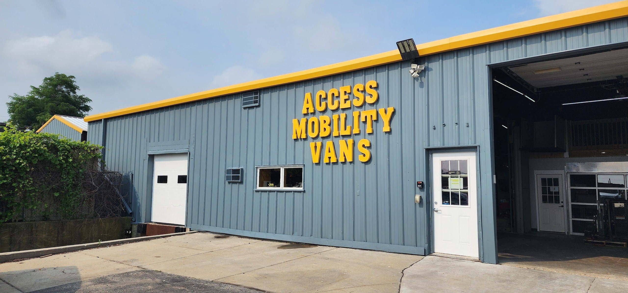 A gray metal building with yellow trim and large yellow letters reading Access Mobility Vans—offering Mobility Solutions Cudahy—above two white doors, one a garage door and the other a standard entry. The sky is partly cloudy.