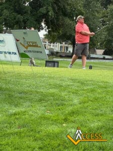A person in a pink shirt and cap swings a golf club on a green course. Nearby, signs display ACCESS branding and sponsor logos. Trees and buildings are visible in the background.