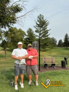 Two men stand on a golf course near a tee marker and hole sign, holding golf clubs. They are smiling, with trees and a bench in the background. The Access logo is in the lower right corner.