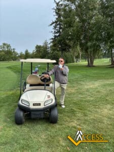A person stands next to a golf cart on a grassy golf course, surrounded by trees. The person is wearing sunglasses and appears to be eating or drinking. The Access logo is in the bottom right corner.