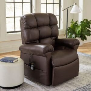 A plush dark brown Cloud Medium/Large Lift Chair by Golden Technologies with a remote in the side pocket sits near a window, next to a white stool topped with a book; a green plant is visible in the background.