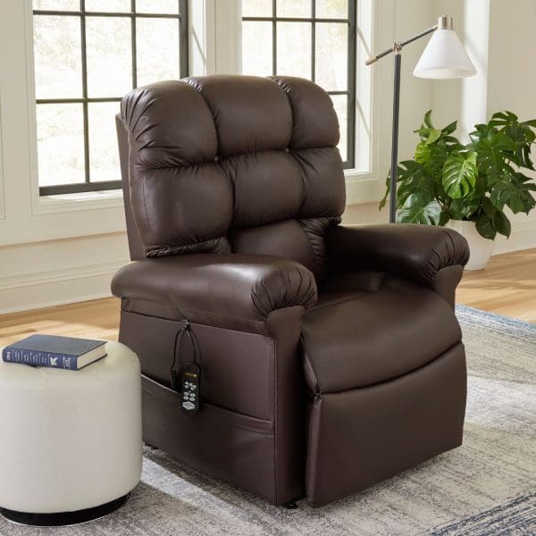 A plush dark brown Cloud Medium/Large Lift Chair by Golden Technologies with a remote in the side pocket sits near a window, next to a white stool topped with a book; a green plant is visible in the background.