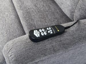 A black remote with white buttons, labeled Up, Preset, and arrows, rests on the arm of a gray Comforter with MaxiComfort Tall Lift Chair by Golden Technologies. The remote is wired to the lift chair.