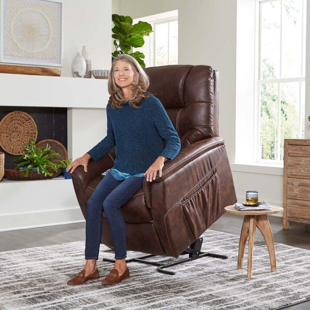 An older woman with long gray hair smiles while relaxing in a Dione Large - Lift Chair by Golden Technologies, a brown leather recliner, in a bright living room with large windows, a patterned rug, and modern decor.