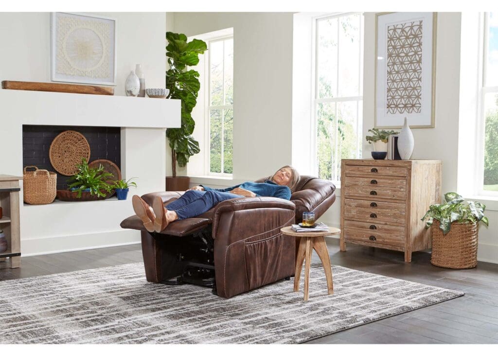 A person relaxes on a Dione Medium - Lift Chair by Golden Technologies in a modern living room with large windows, light wood furniture, a gray patterned rug, and green plants.
